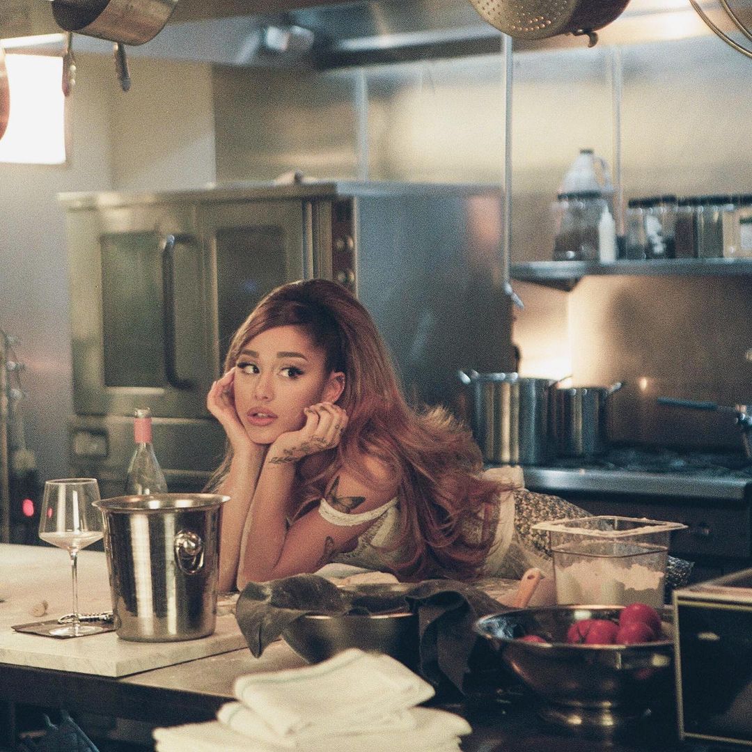 A photo showing Ariana Grande leaning on her kitchen counter and there's a bottle of wine and wine glass in front of her.