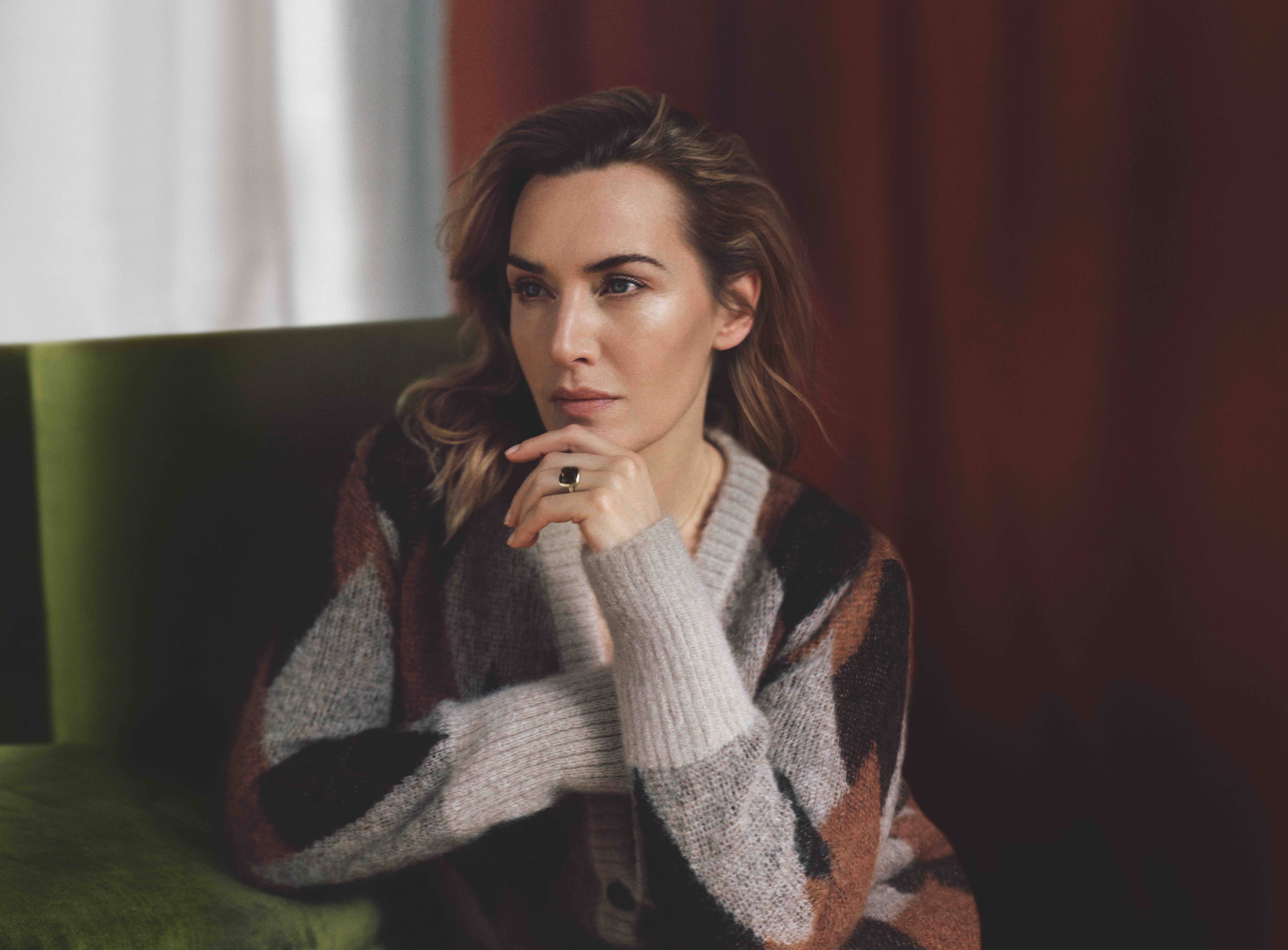 A photo showing Kate Winslet adorned in a multicolored sweatshirt, sitting with her hand on her jawline.