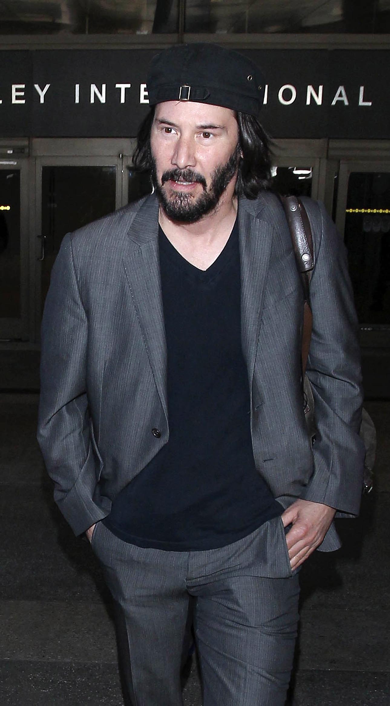 A photo showing Keanu Reeves in a gray suot and pant, with a hand luggage.