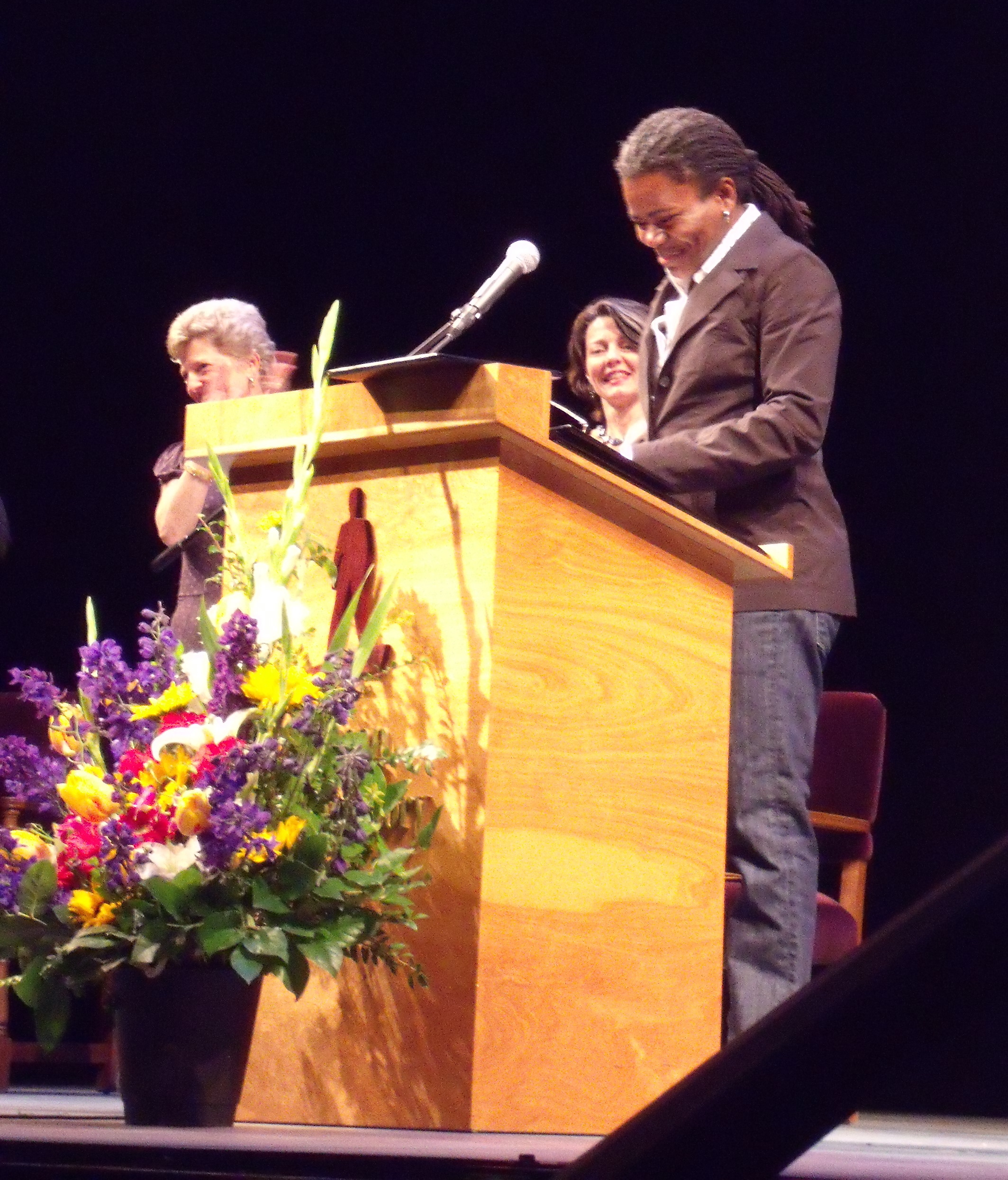 A photo showing Tracy Chapman, giving a speech at an event and she has on a black suit with denim pants on.