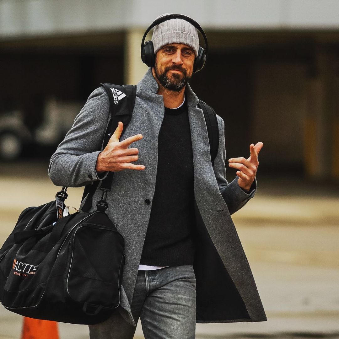 Aaron Rodgers looks stylish in this gray jacket on a black plain shirt and denim pant, with a gray color head-warmer to match and a luggage.