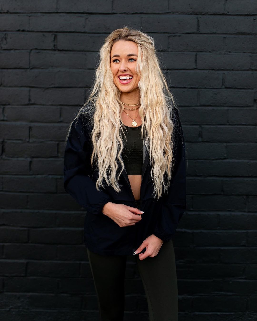 A photo showing Heather Martin in a sweatshirt and pant with top-bra, while posing for a photo in a black brickwall background.