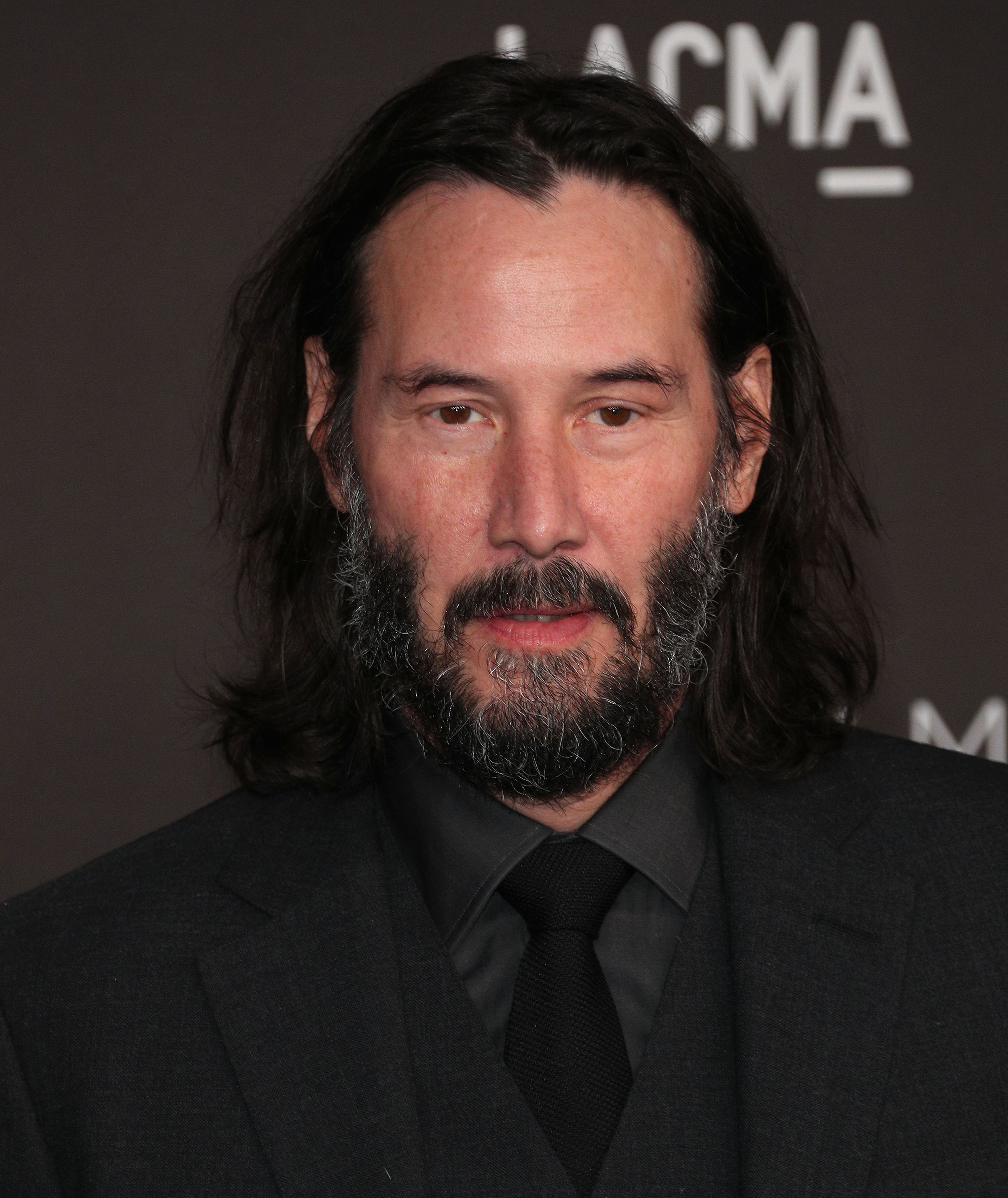 A photo showing Keanu Reeves rocking a black suit with a black inner T-shirt and tie.