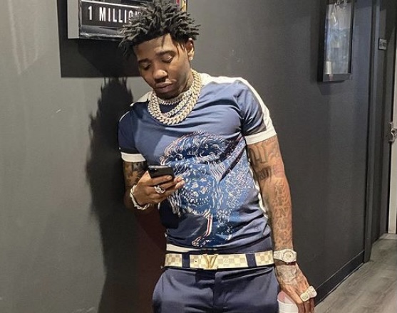 Lucci checking his phone while showing his Versace t-shirt and Louis Vuitton belt.