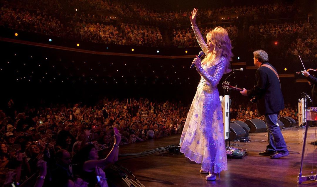 Dolly Parton, in a white dress on a huge stage, waves to a hyped crowd consisted of thousands of fans.