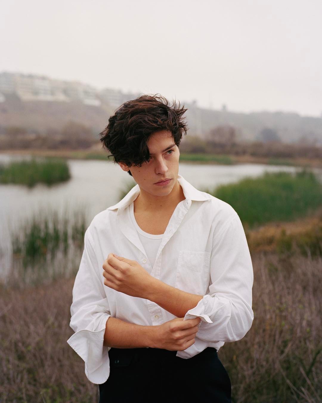 Cole Sprouse looking dazzling in a beautiful white shirt and black pants, near a shrub-filled place.
