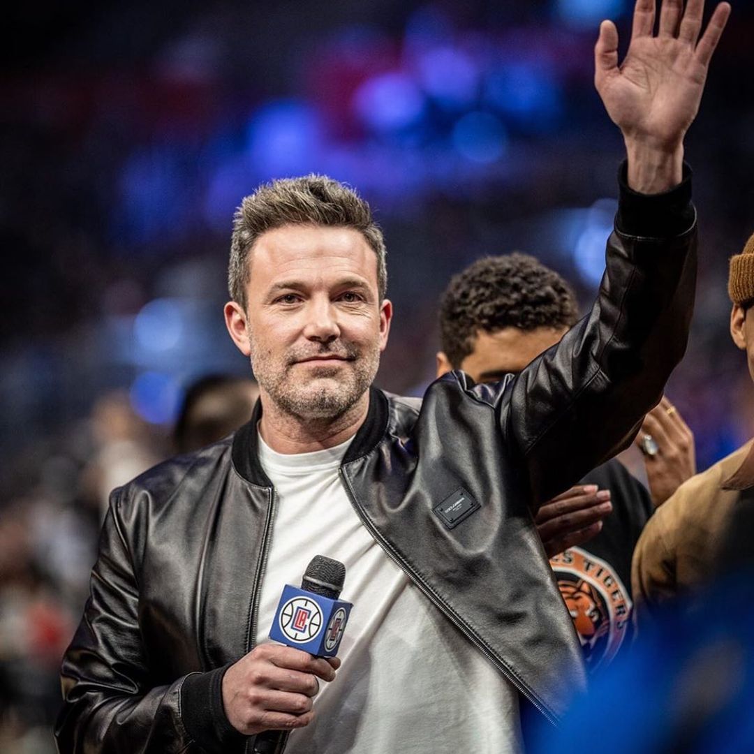 A photo of Ben Affleck in a black leather jacket over a plain white round-neck shirt, and he's holding a microphone in one hand and the other raised to acknowledge fans.