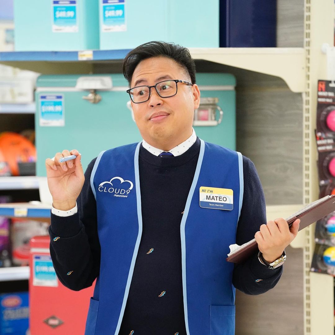 A photo of Nico Santos from a scene in 'Superstores' and it shows him in a Cloud 9 uniform with a note-pan and pen in his hands.