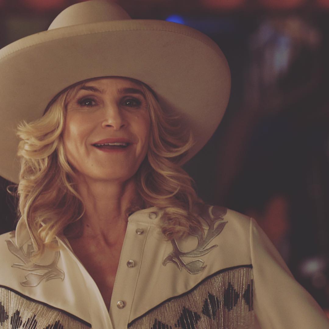 A photo of Kyra Sedgwick sporting a white cow-girl hat and white T-shirt with black designs in the chest area.