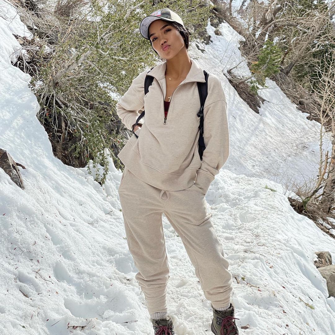 A photo showing Jhené Aiko in a matching sweatshirt and pant with green boots and a black backpack posing for a photo in the mountains.