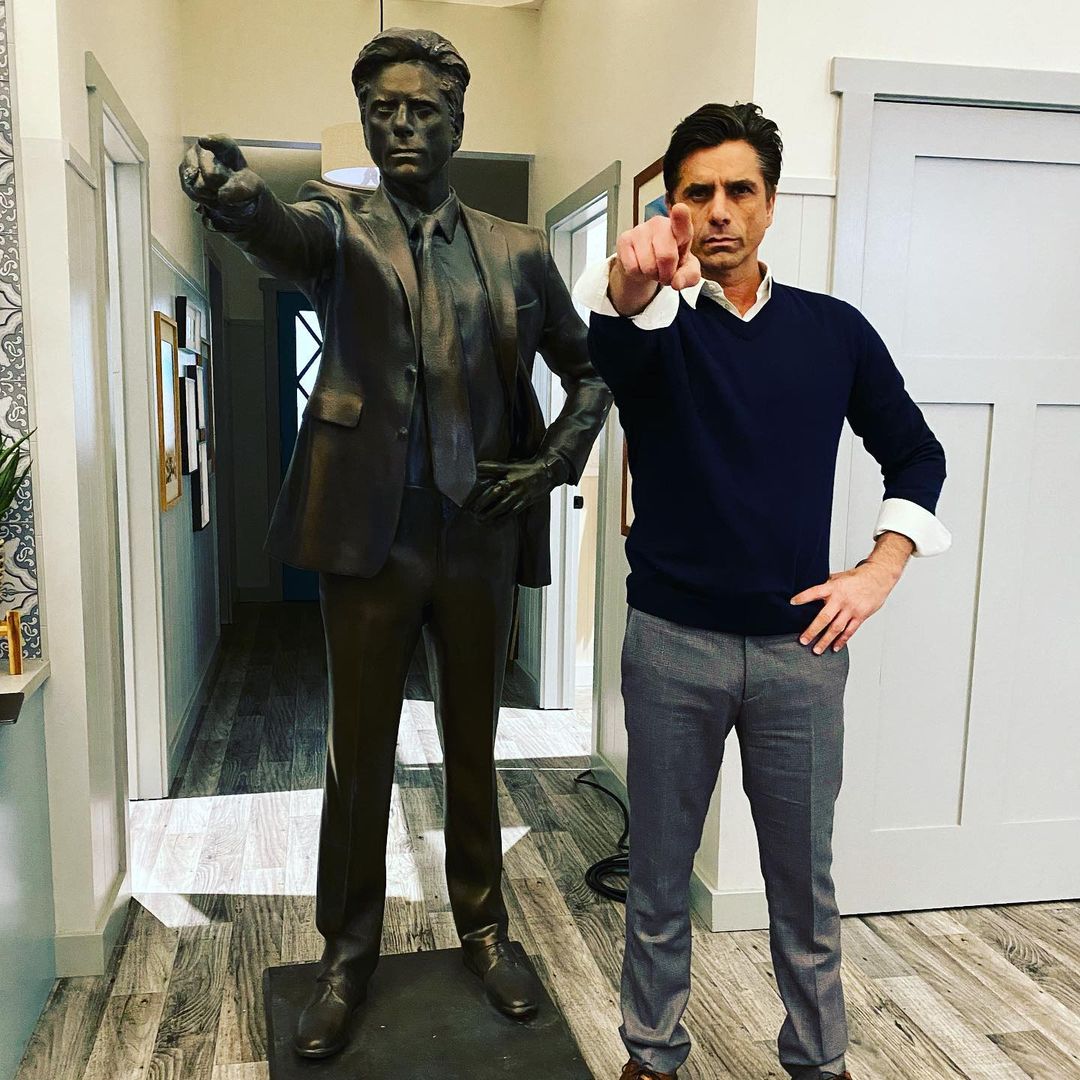 A photo showing John Stamos standing beside a statue of himself with a hand pose.