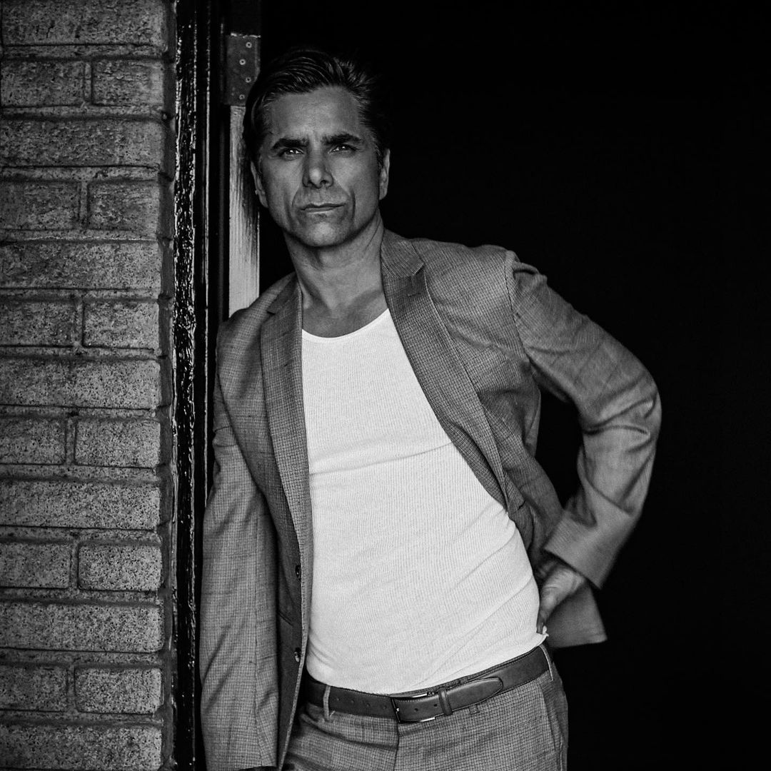 A black and white themed photo showing John Stamos in a gray suit and pant.