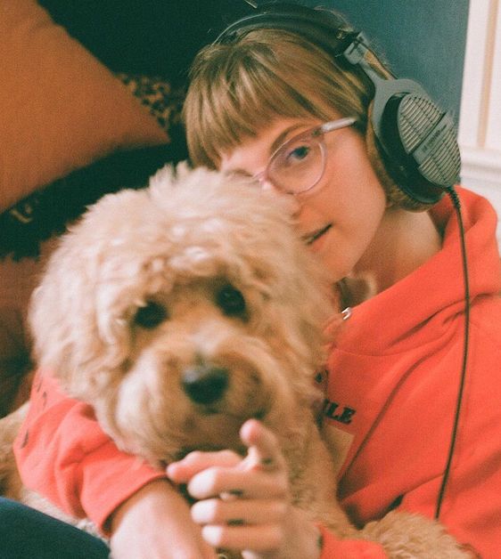 Hayley Williams wears a bright orange sweatshirt as she takes a photo with her dog as she points into the camera.