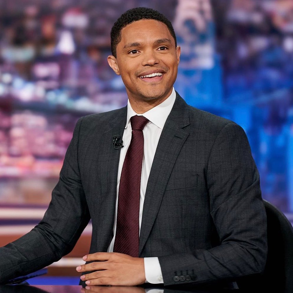 Trevor Noah in a black suit, white inner shirt and a brown tie.