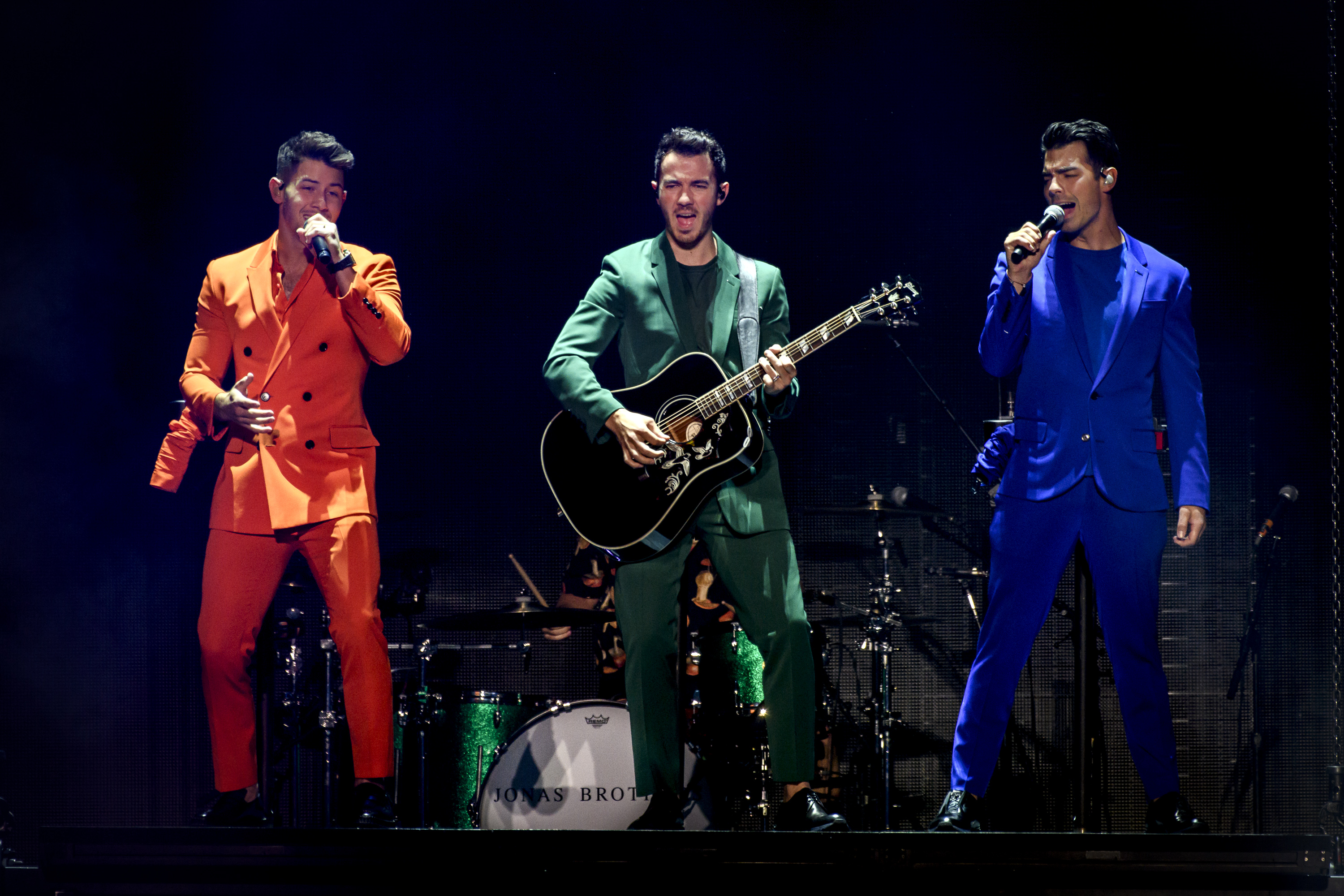 A photo of the Jonas Brothers performing on stage in multi-colored suits.