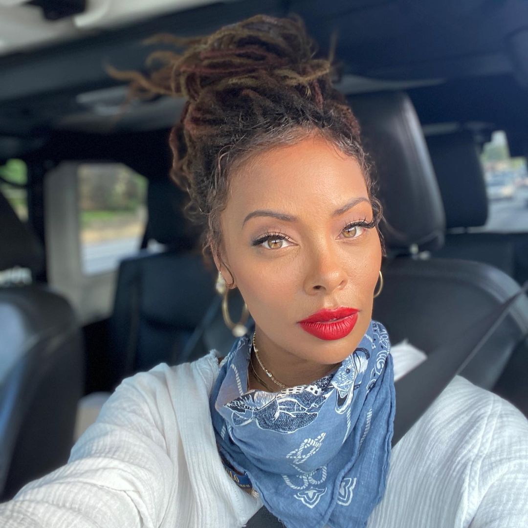 A photo of Eva Marcille in the driver's seat of a vehicle wearing a white longsleeve outfit with a blue scarf around her neck.