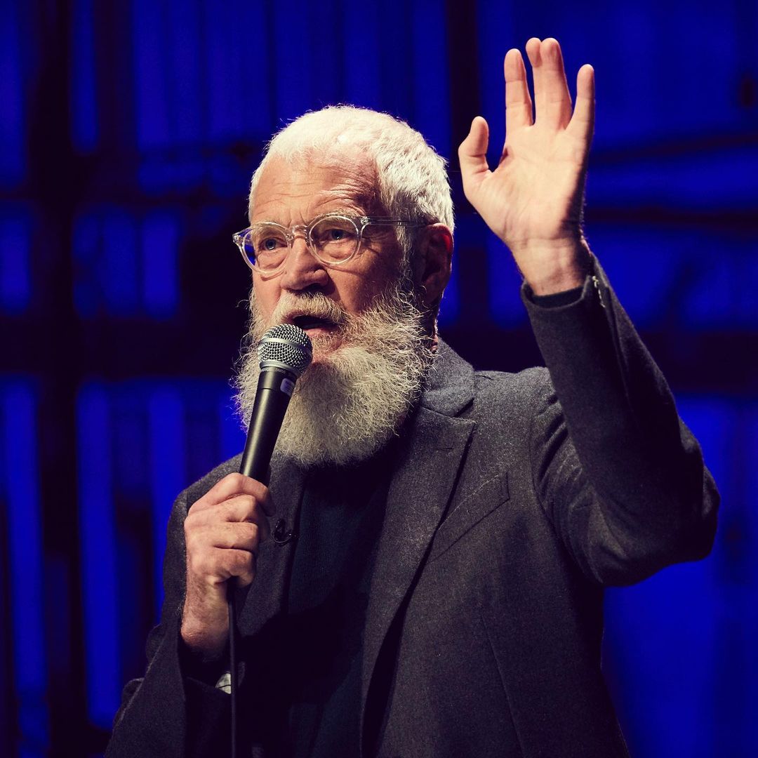 David Letterman looks amazing in this dark gray suit, while giving a speech with his hand raised in the air.