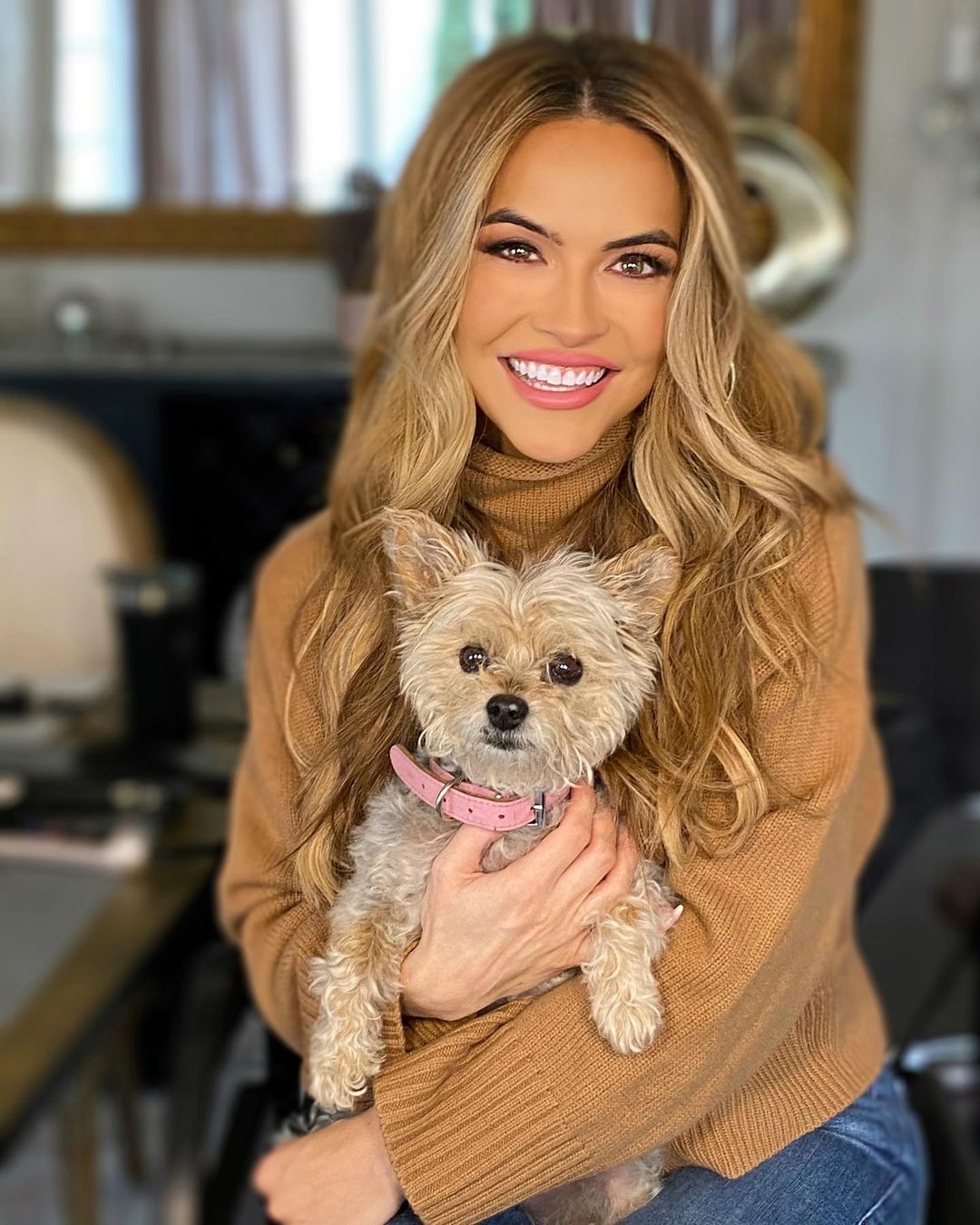 A photo showing Chrishell Stause in a brown sweater and blue denim pant, holding on to her beautiful puppy.