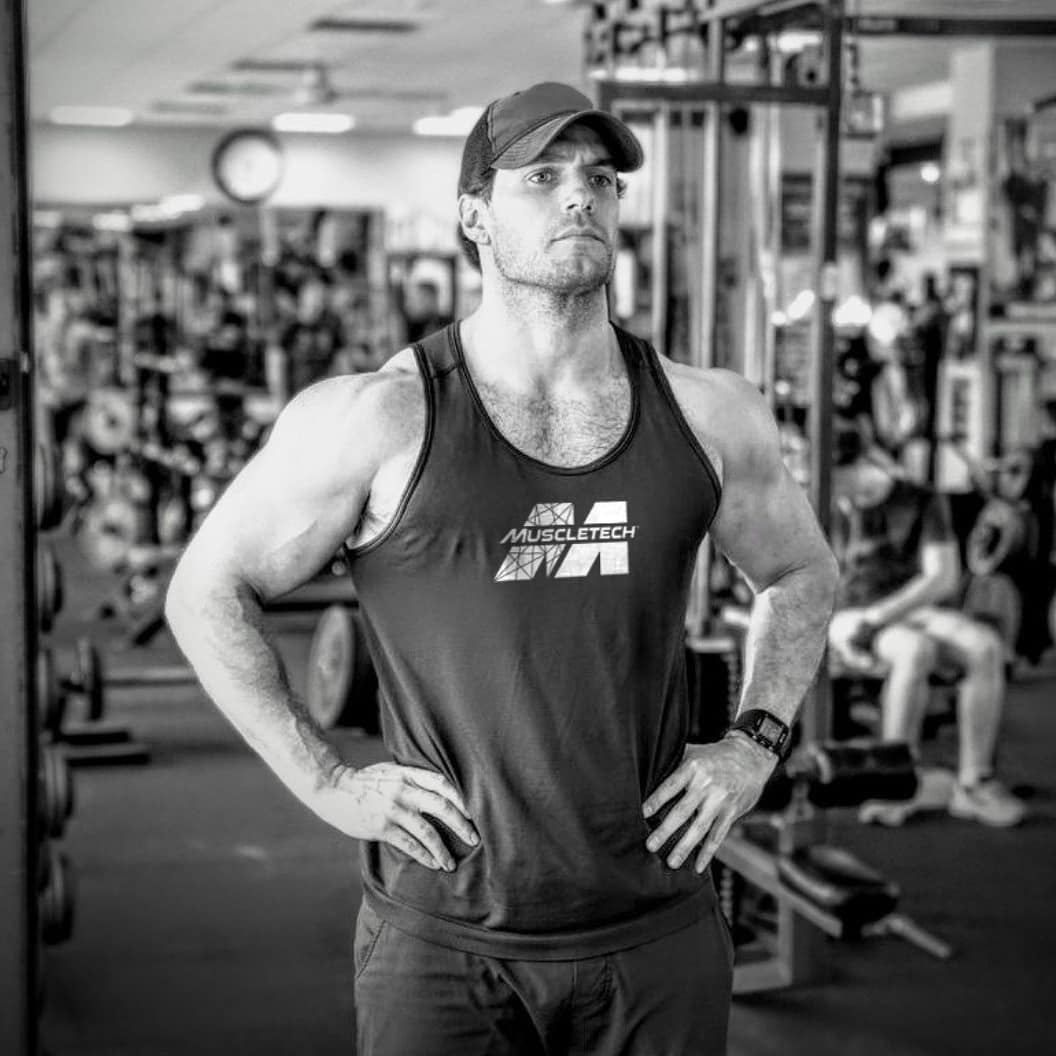 A black and white themed photo showing Henry Cavill posing for the camera with a straight expression on his face at the gym.