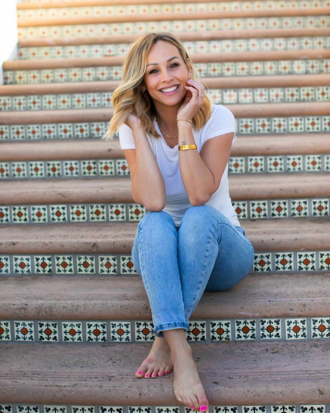 A photo showing Clare Crawley sitting on a flight of stairs bare-footed in a white blouse and blue denim pant.