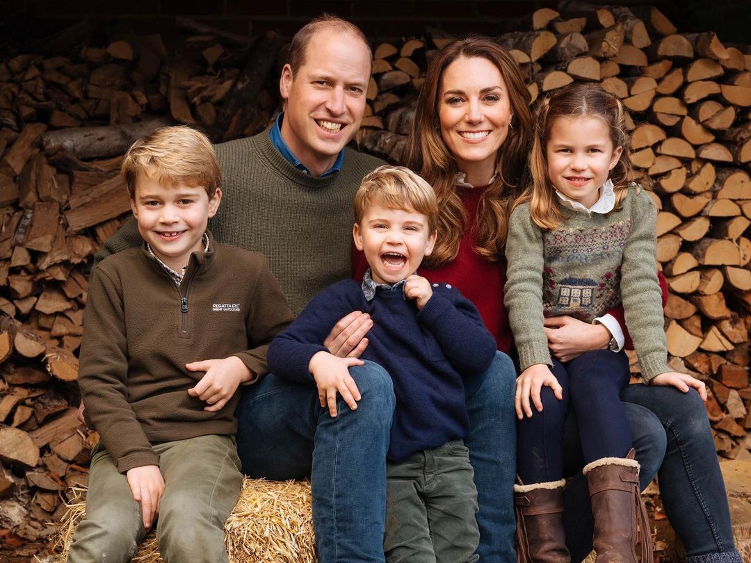 A family photo featuring Kate Middleton and Prince William, with their beautiful kids, in front of piled woods.