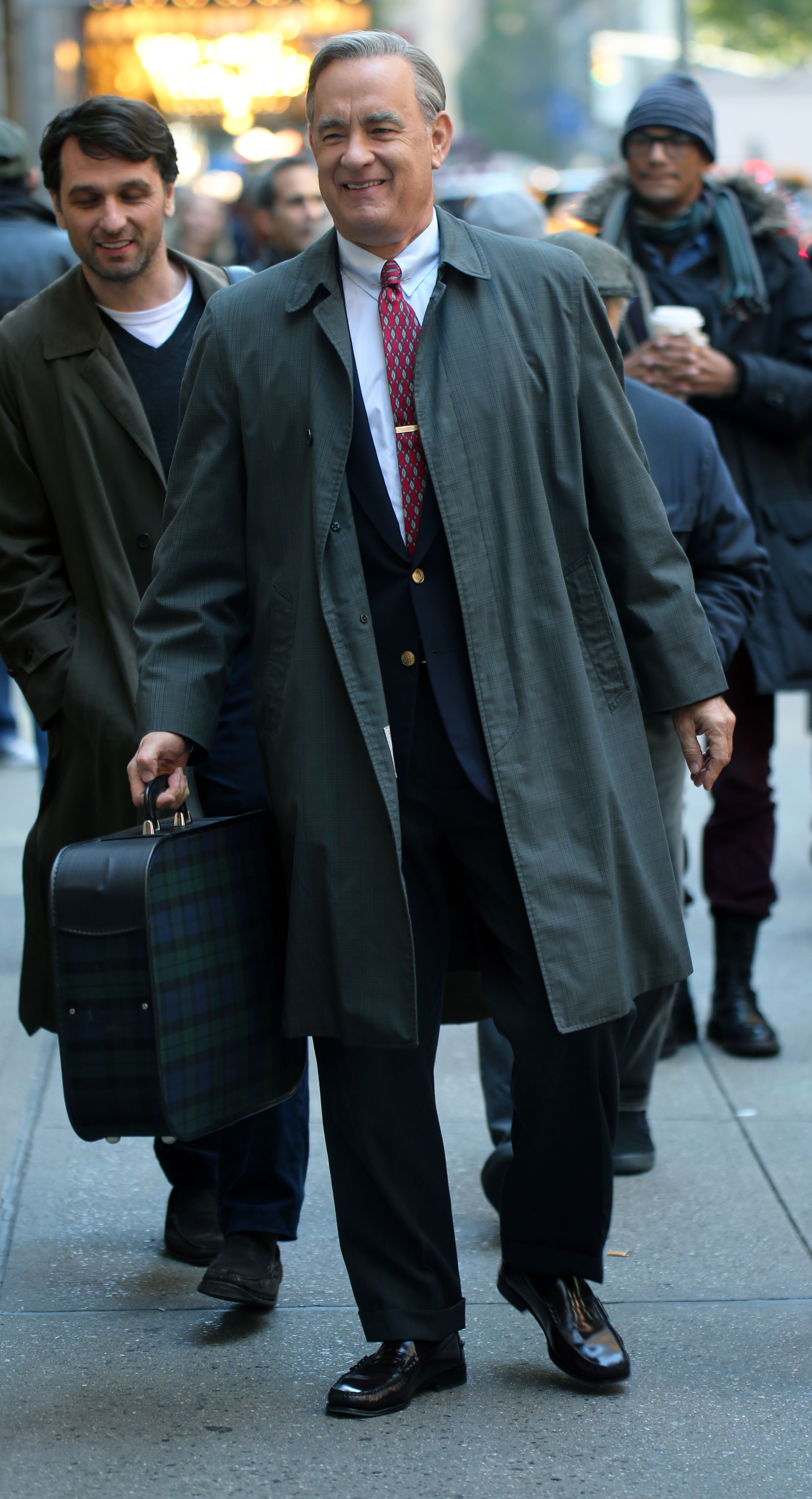 A photo showing Tom Hanks on the streets  in a long jacket and pant, holding a large briefcase.