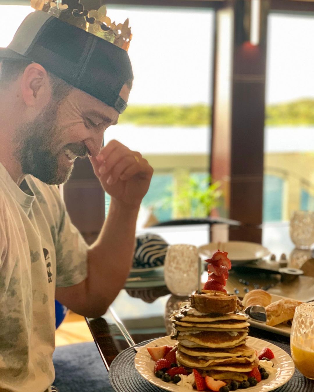 A picture of Justin Timberlake staring at a plate of piled pancakes, with strawberries and blueberries around it, with a smile on his face.