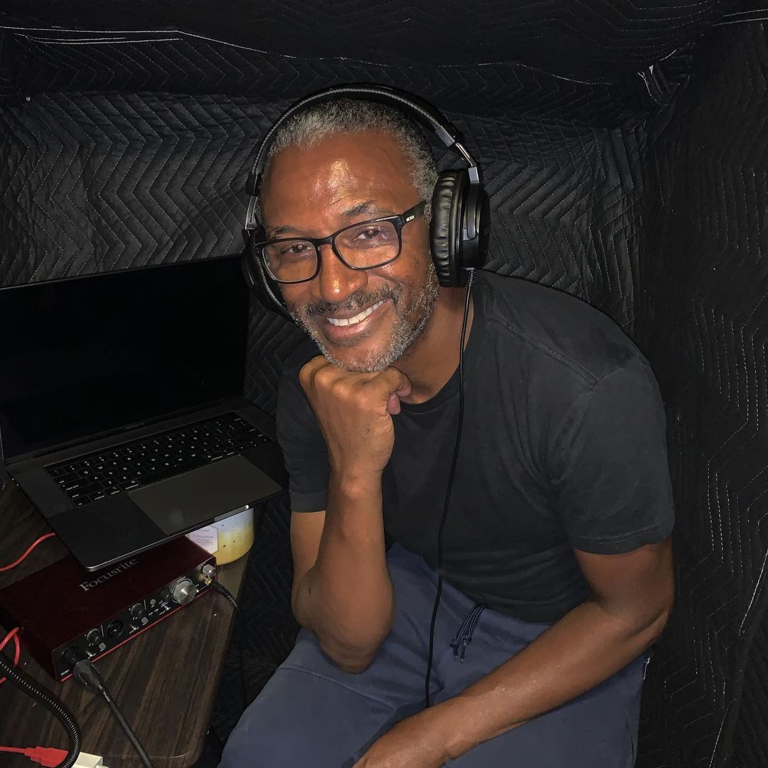 A photo of Tommy Davidson sporting a casual shirt and joggers, while listening to music with a black headset that's connected to a laptop.
