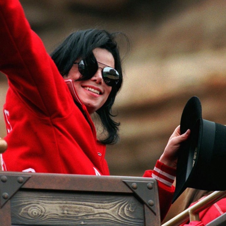 A picture of Michael Jackson wearing a red jacket while waving with a beautiful smile on his face.