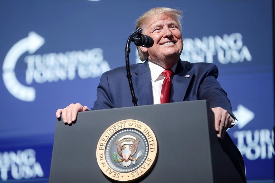 A photo showing President Donald Trump speaking from a podium, in a blue suit, with a white inner T-shirt and red tie.