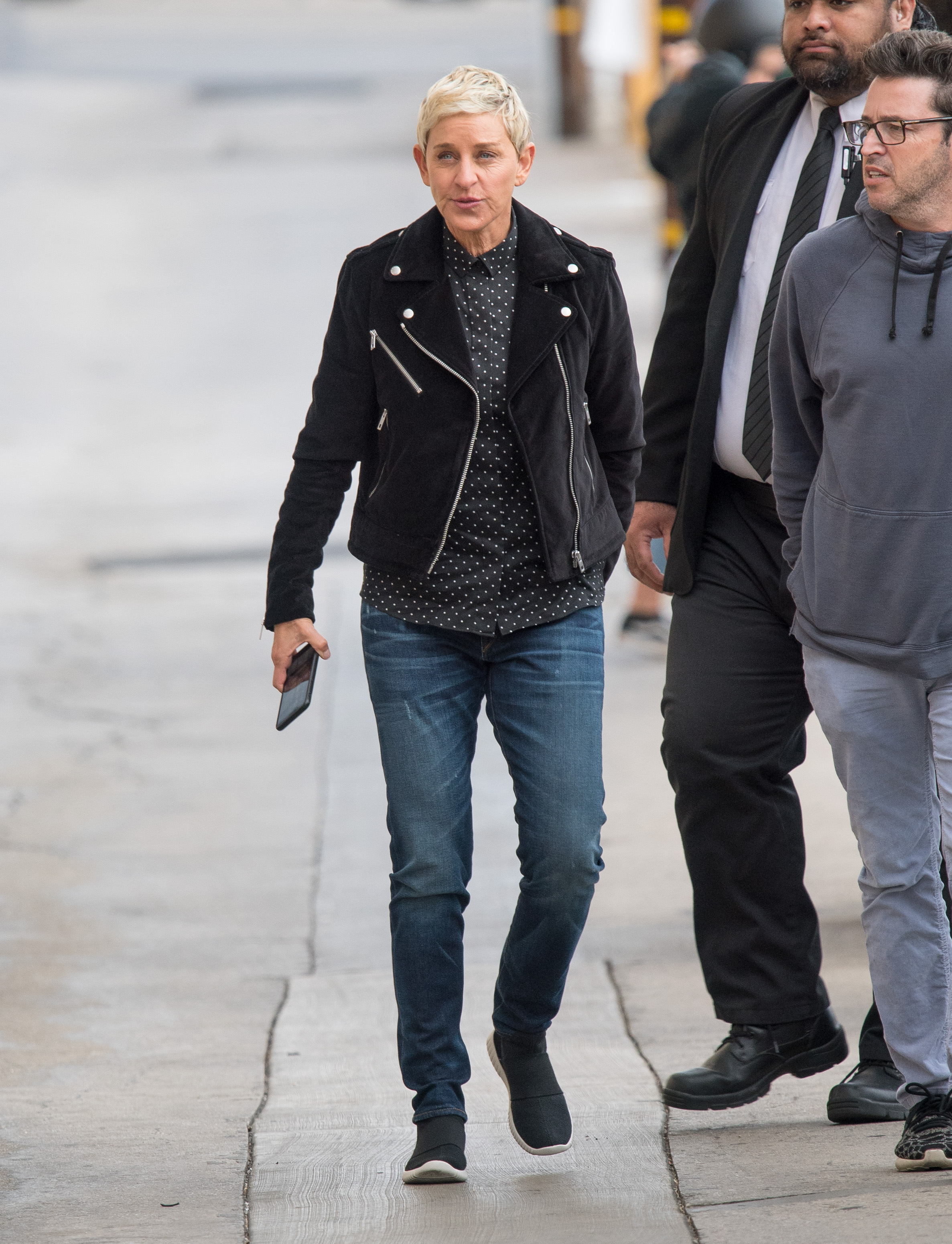 Ellen DeGeneres captured on camera in public in a blue denim pant, black sneakers , a black jacket over a polka dot design shirt.