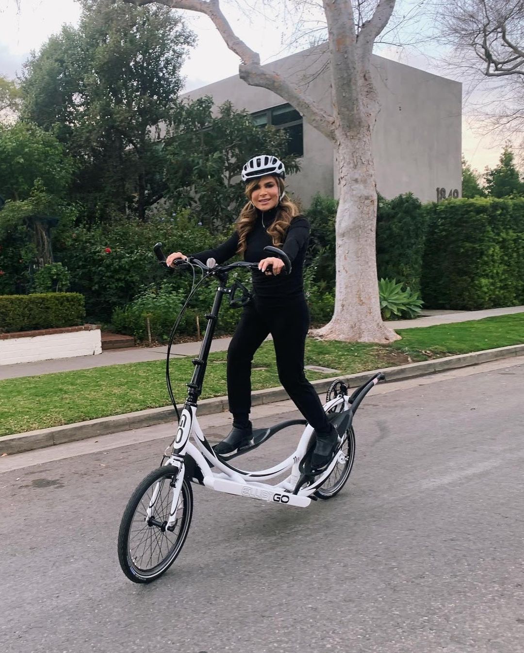 A photo of Paula Abdul in a black longsleeve and pant, riding a scooter on a street.