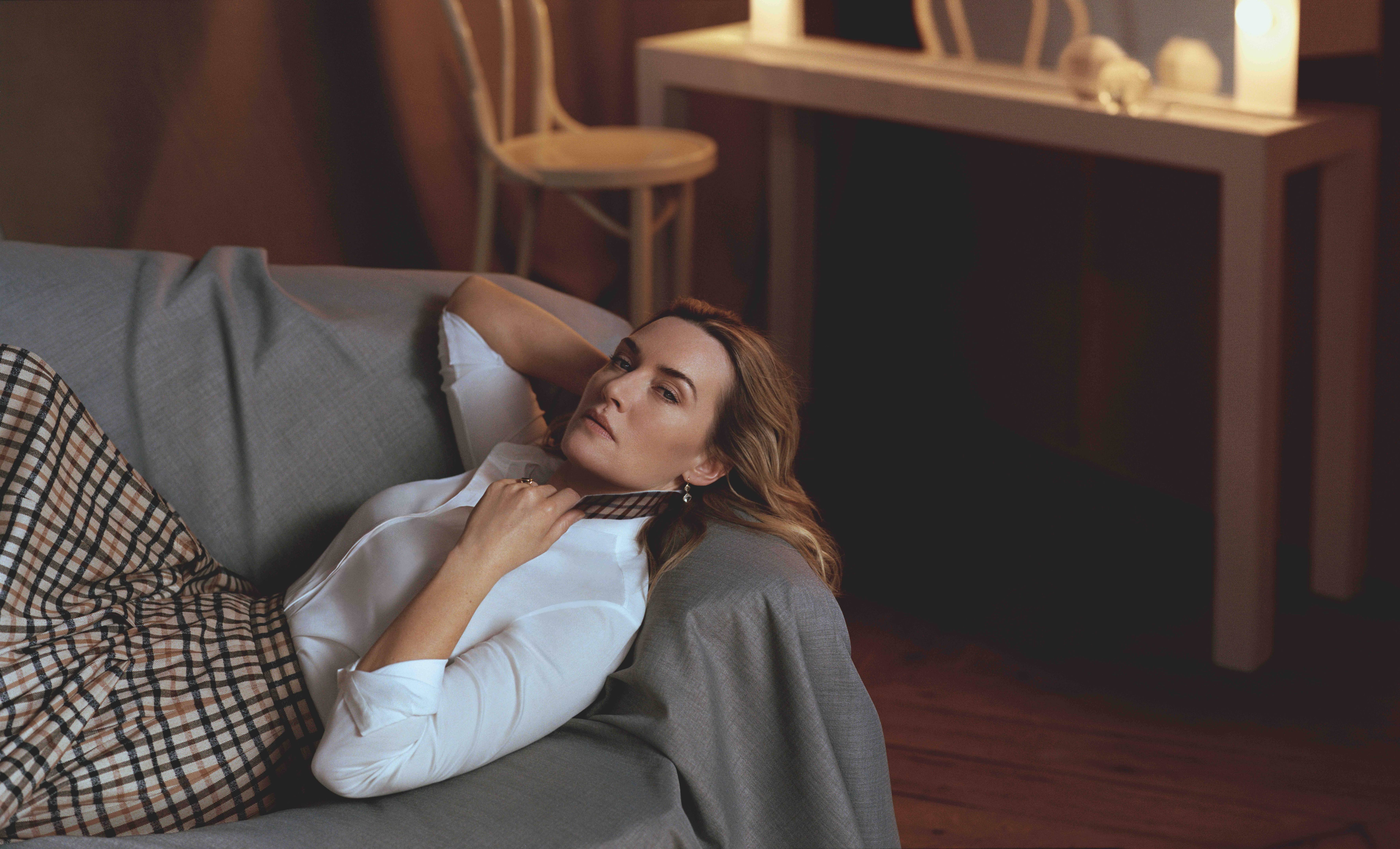 A photo showing Kate Winslet in a white T-shirt and check material skirt, and she's laid down on a sofa covered in gray linen.