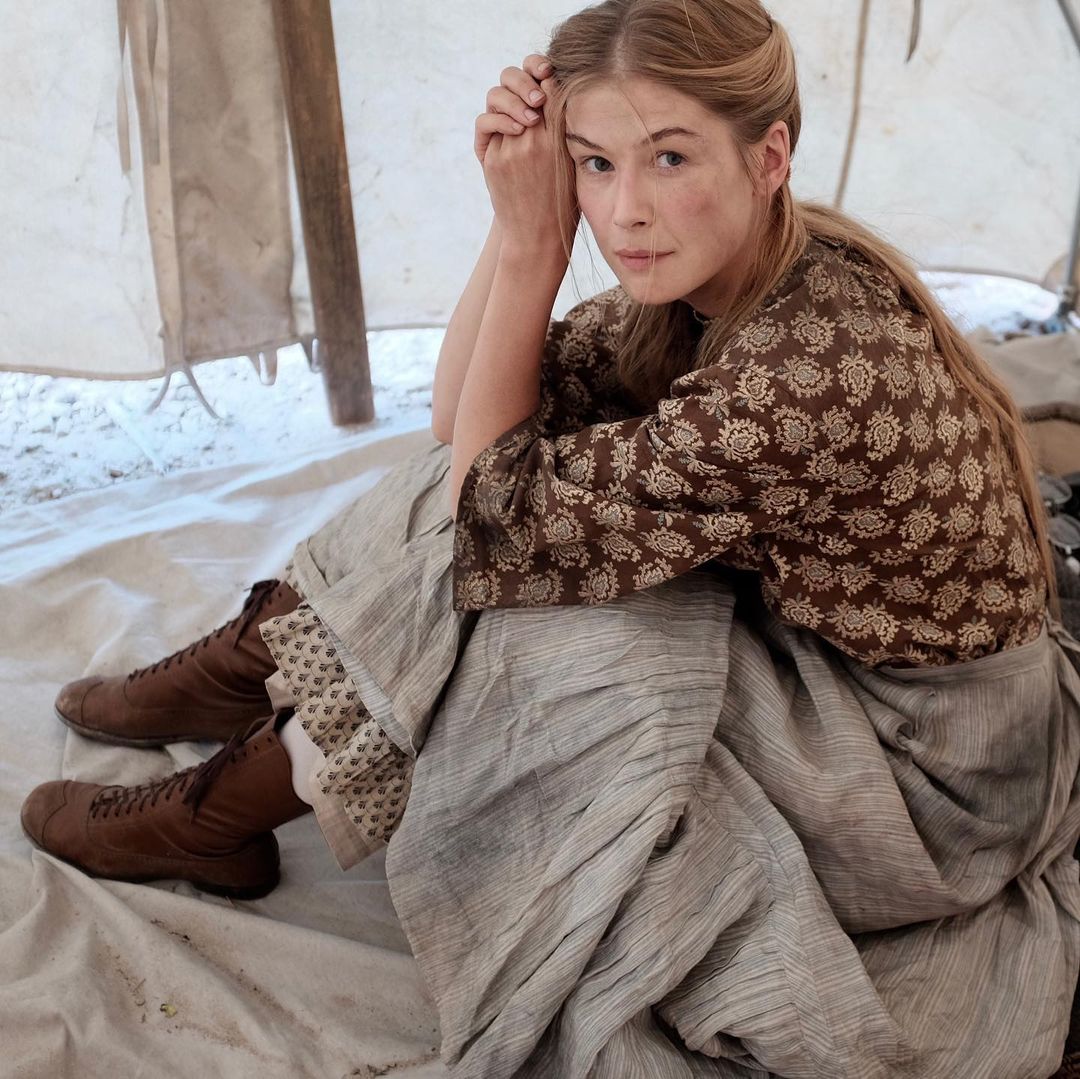 A photo of Rosamund Pike from a movie scene, sporting a pair of brown boots, flower-design blouse and pleated skirt.