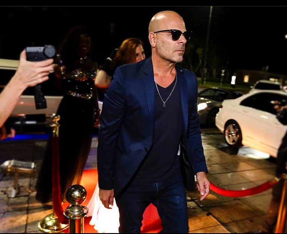 A photo of Bruce Willis looking handsome in a blue suit with a black inner shirt matced with a pair of jeans and sunshades.