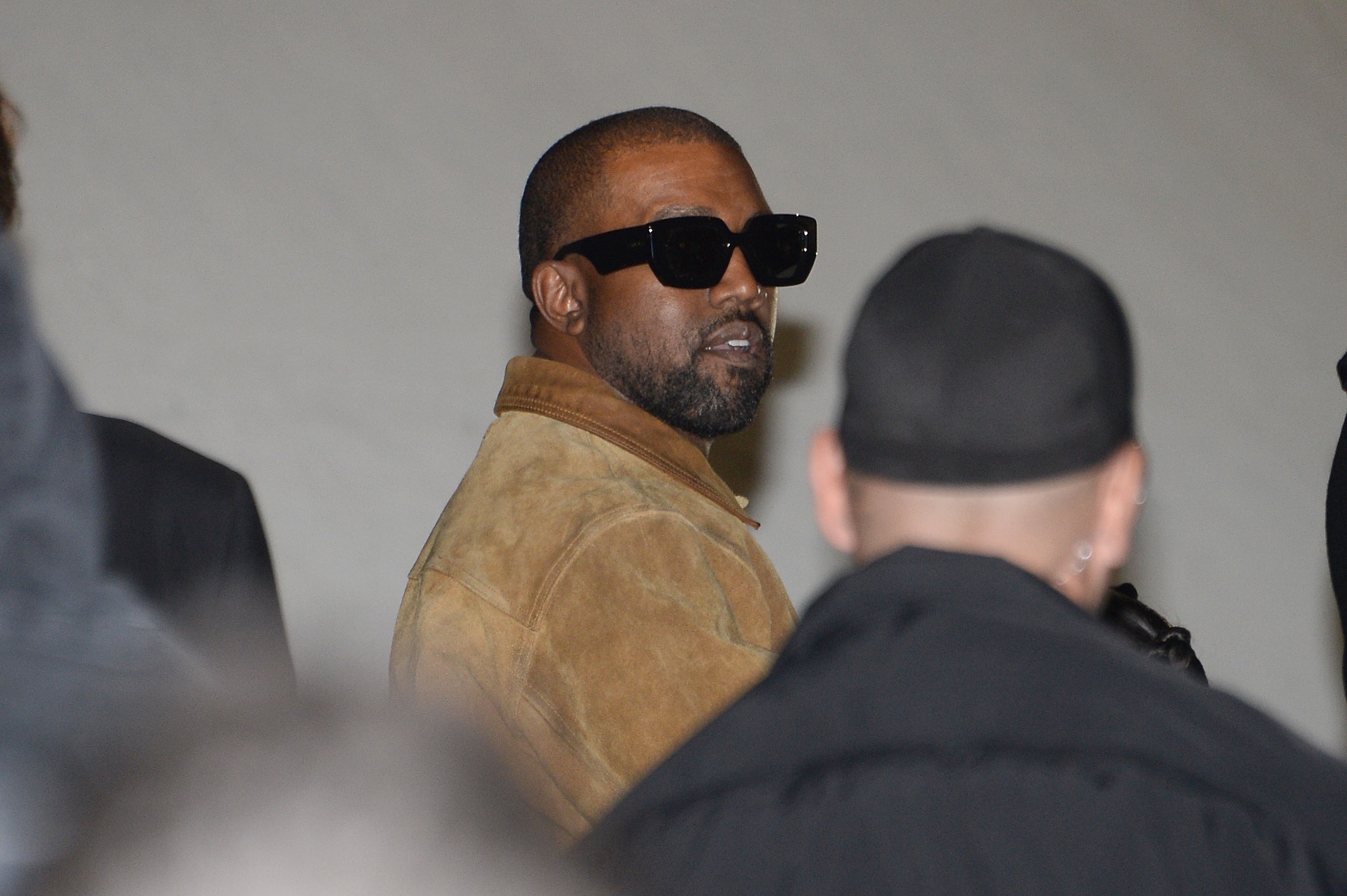 A photo showing Kanye West dressed in a brown jacket and black shades, at a fashion show in Paris.