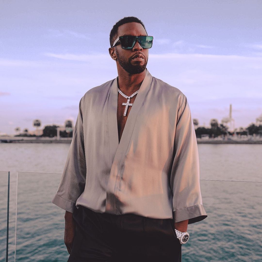 A photo of Diddy in a brown designer shirt with black pants and a silver chain around his neck with sunshades and a beautiful ocean view behind him.