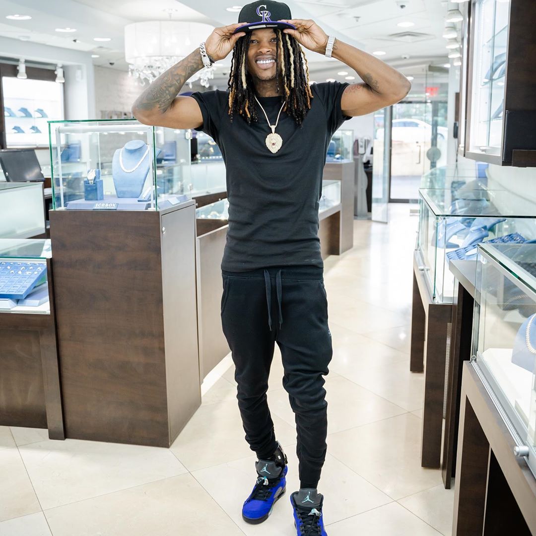 A photo of King Von at a jewelry store, sporting a black pant and shirt, with a huge gold necklace.