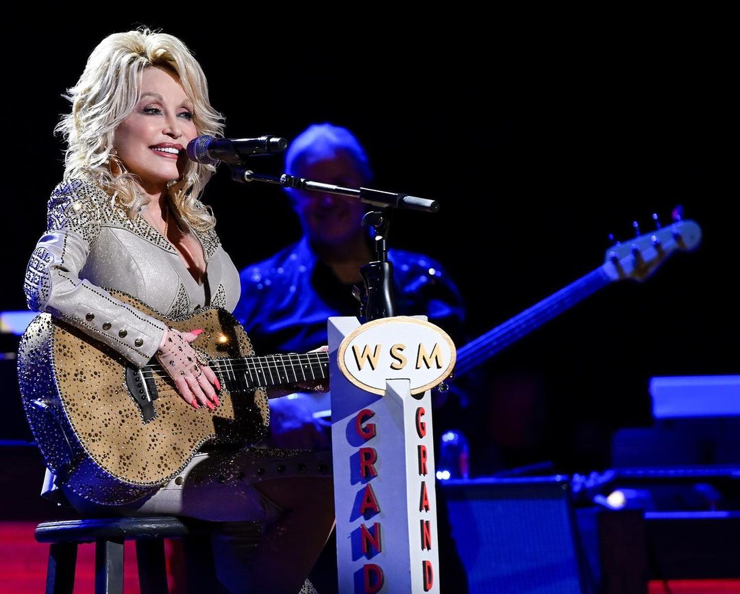 A photo of Dolly Parton taken in motion on a stage, as she prepares to sing with her beautiful guitar.