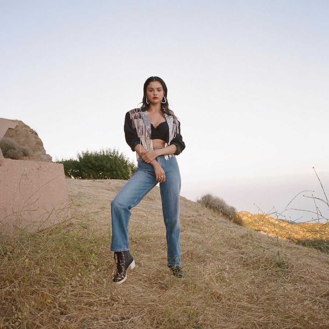 A photo of Selena Gomez in a field area, dressed in a jacket and denim pant. 