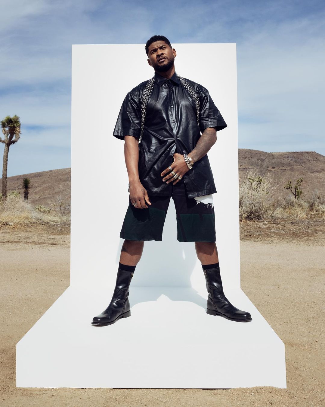 A photo showing Usher standing on a white color platform in a black leather outfit and shorts to match. 