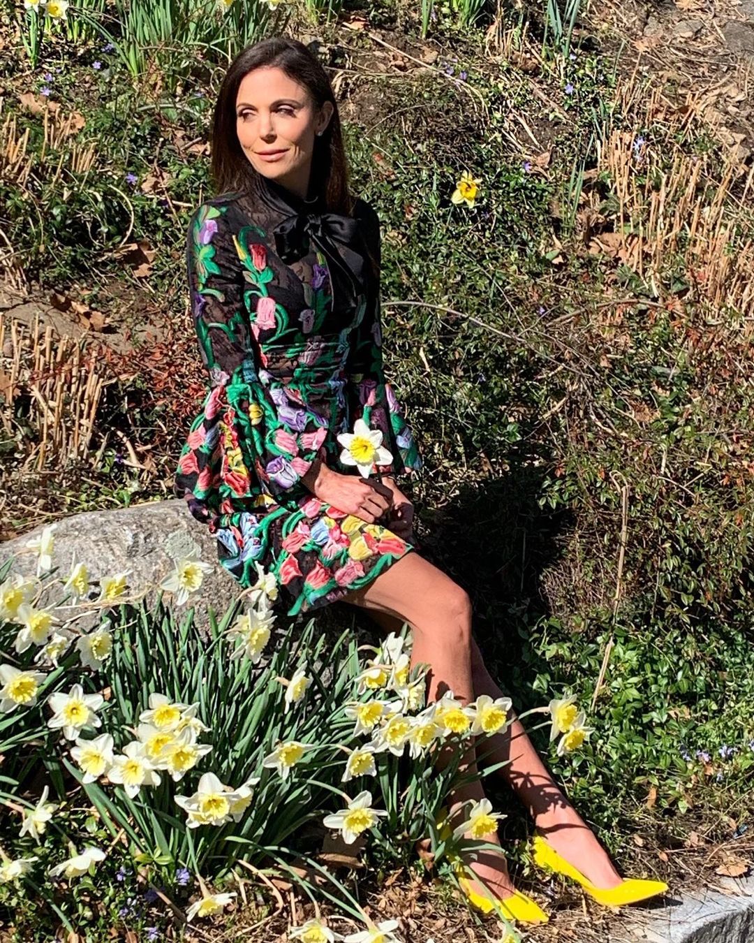 A photo showing Bethenny Frankel posing in a flower garden, filled with sunflowers and she has on a multicolored flowery design dress and yellow color pumps to match.