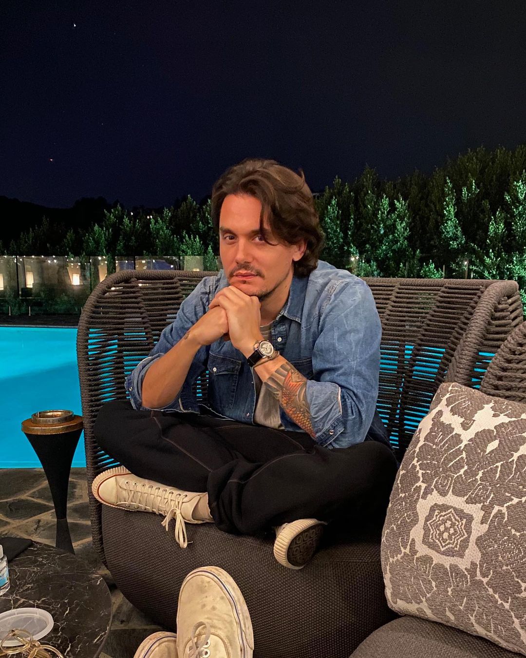 A photo showing John Mayer sitting on a couch by a pool, with his legs tucked in and his hands rested on his lower chin.