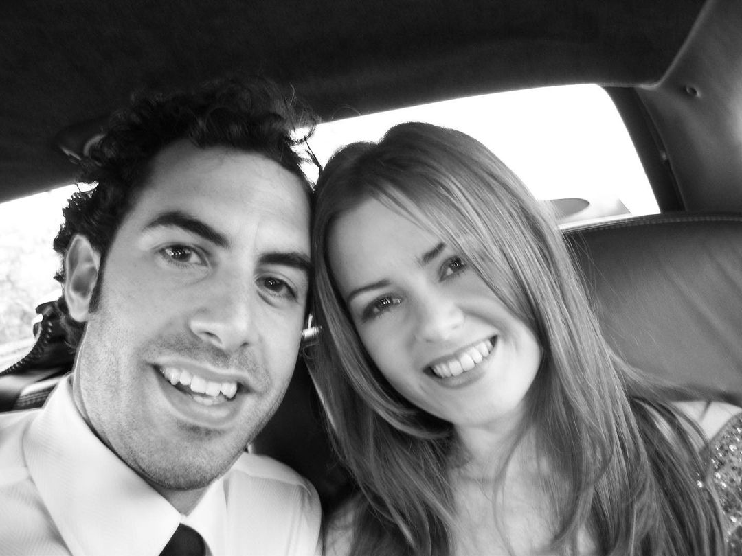 A black and white themed photo of Isla Fisher and Sacha Baron Cohen in the back seat of a vehicle.