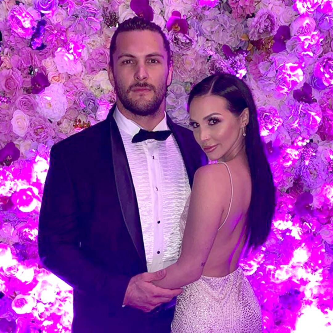 Scheana Shay wears a backless dress in front of a wall of flowers.