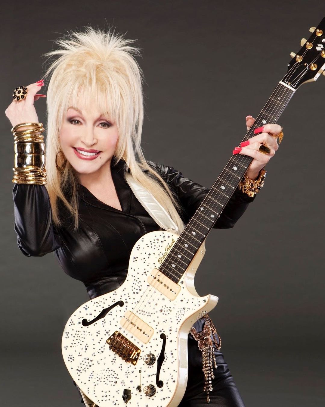 A photo showing Dolly Parton with a guitar in her hand, smiling beautifully as she poses for the camera.