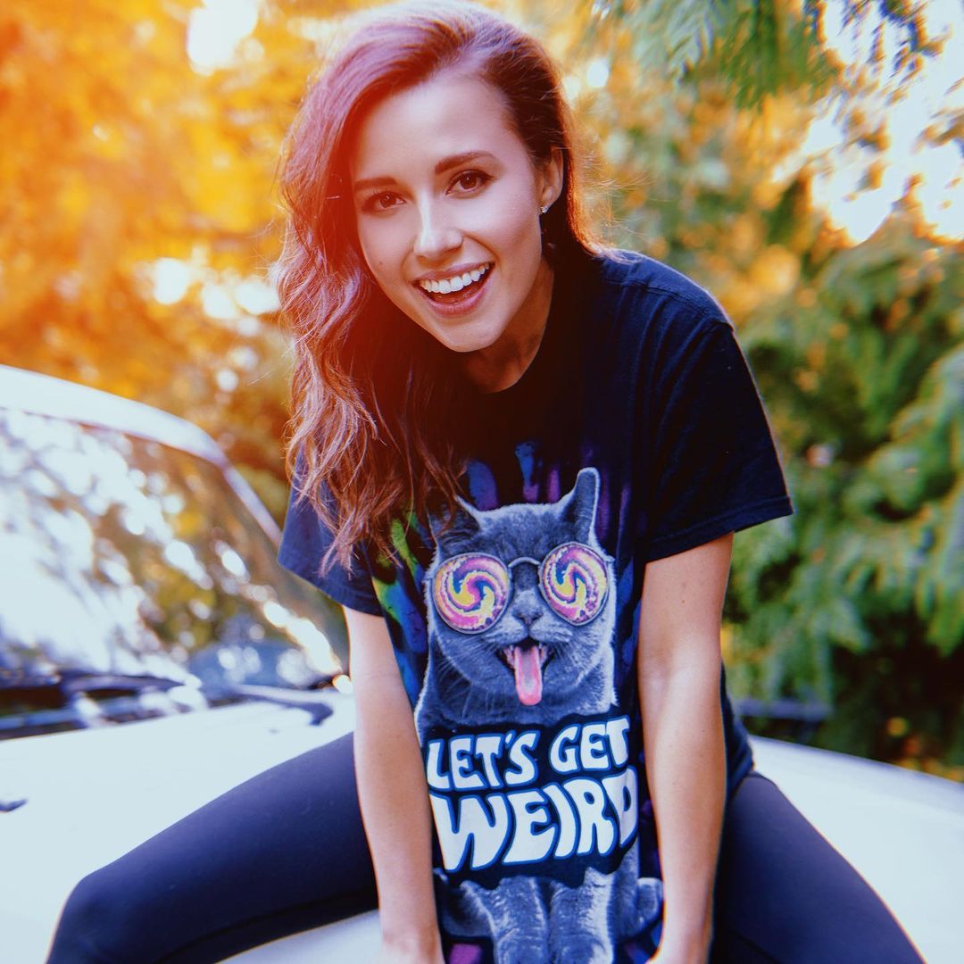 An amazing photo of Katie Thurston sitting on the hood of a car, rocking a cat-design shirt and black pant.