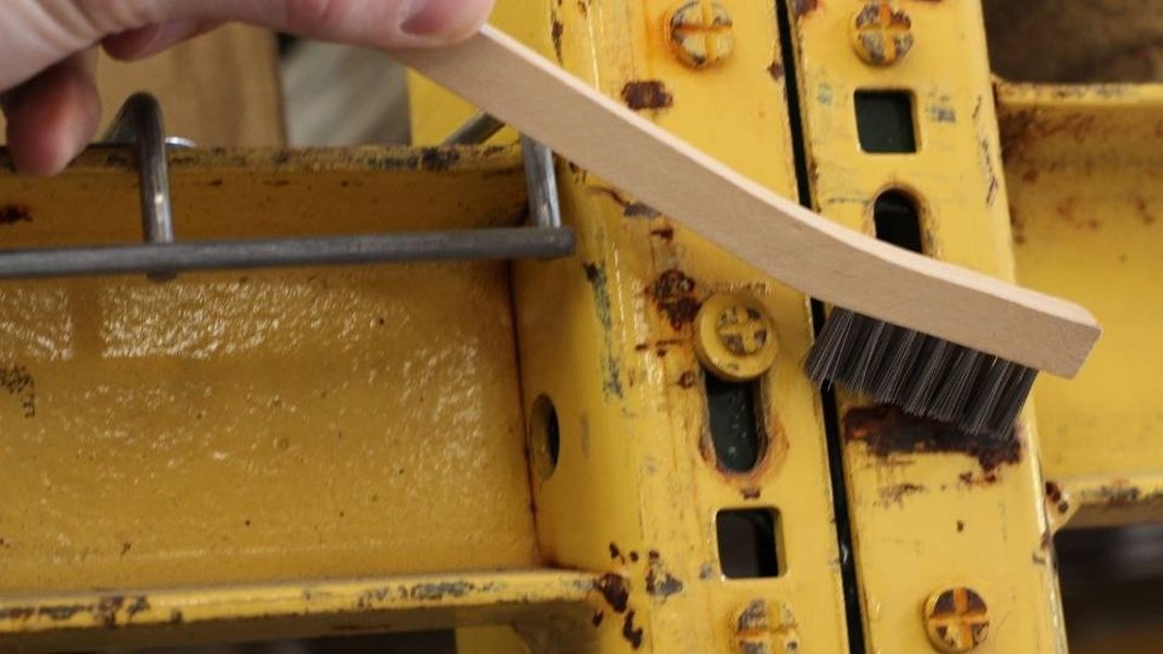 A person using a handheld wire brush to remove rust from a metal surface.