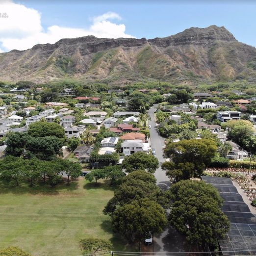 This aerial view shows a lush residential neighborhood of single-family homes nestled at the base of the massive, rugged slopes of Diamond Head crater in Hawaii.A wide green park spreads across the foreground on the left,balanced by a shaded plant nursery