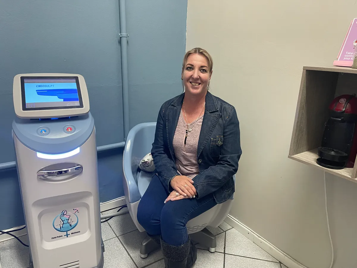 A woman sits smiling in a light blue pelvic floor therapy chair next to the Embosulpt PT machine.
